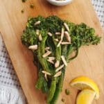 A close up of a rectangular wooden board of food with broccoli, lemon slices and a bowl of slivered almonds on it against a white and grey table cloth.