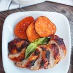 A white plate on a brown wooden countertop. On the plate is grilled a sliced grilled chicken breast and yam slices garnished with a sprig of basil. In the top of the photo, there is a bowl of rice on top of a white cloth napkin.