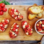 Caprese Bruschetta on a wooden cutting board with various ingredients spread around on a grey counter top.