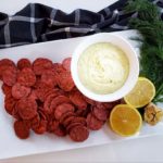 Beet crackers arranged on a plate with white bean dip (in a bowl), lemon, garlic and dill as a garnish. The white plate is on top of a navy blue checkered cloth napkin against a white background.