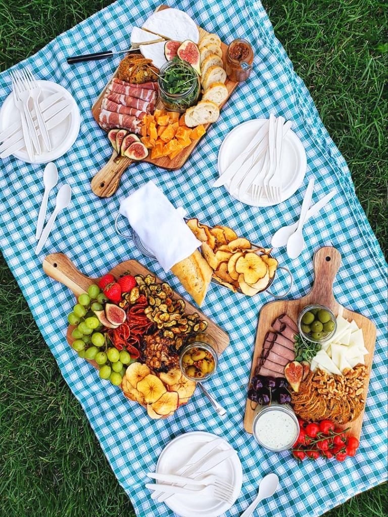 Charcuterie boards on a blue plaid picnic blanket with a grass background. Around the three boards are plates with compostable cutlery, and a plate that has a baguette and apple crisps on it.