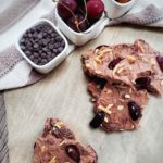 Cherry and almond chocolate frozen yogurt bark on a counter with parchment paper. In the background, there are three separate small white square bowls. One bowl has chocolate chips, the second cherries and the third has almonds in it.