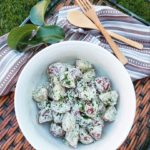 A creamy potato salad garnished with herbs on a wicker tray with a cloth napkin and wooden utensils on a grass background.