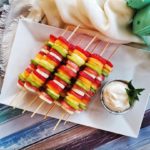 Mixed skewered fruit on a white plate with a container of a white "cheesecake" dip against a multicoloured wood background.