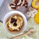 Peach and mango Nana ice cream topped with candied pecans surrounded by ingredients and a wooden spoon on a wooden counter top that is half covered by a white tablecloth with a green design on it.