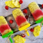 Raspberry and pineapple popsicles in plastic holders with twine tied around the handles. The popsicles are in a metal tray on ice, raspberries and pineapples. The metal tray sits on top of a concrete counter top.