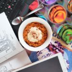 A top-down view of "Carrot Cake Oatmeal" in a white bowl against a messy desk background. On the desk are various papers, textbooks, an iPad, an open laptop and a spoon.