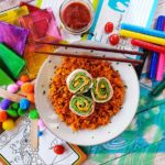 A top-down shot of "Cheesy Green Eggs And Ham Roll-Ups" on a white plate with mahogany-coloured chopsticks on a messy wooden desktop background. On the desk are various papers, markers, stencils and a small mason jar of sauce.