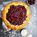 A red cherry our with n open top and golden crust sitting on a wire rack against a stone counter top background that is covered in flour. There is a decorative napkin, bowl of cherries, bowl of flour and a wooden spoon on the counter as well.