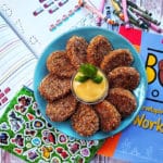 Hidden broccoli chicken nuggets on a blue plate arranged around a yellow sauce garnished with basil leaves. The background is a white wooden table with children's coloring books scattered around the plate.