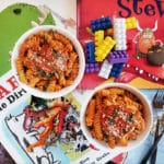 Two bowls of pasta garnished with cheese and herbs on a brick background with children's books scattered around it and two metal forks.