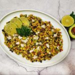 A top-down shot of Summer Corn and Black Bean Salad garnished with parsley leaves and lime slices on a white oval plate on top of a cracked-concrete counter top background. Also on the counter top is a half of an avocado with the pit inside it and half a lime.