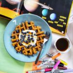 A top-down shot of "Sweet Potato Waffles" on a blue plate with a fork and a small bowl of maple syrup off to the side against a messy desk background. On the desk are various coloured papers, Sharpie markers and an open book about the solar system displaying the planets and some informational text.