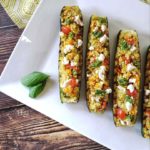 A top-down shot of Zucchini and Tomato Summer Salad Boats on a rectangular white plate garnished with two basil leaves on a wooden background that has a decorative napkin on it as well.