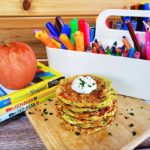 "Zucchini Fritters" garnished with herbs on a wooden cutting board. Surrounding the dish are some decorative books with a red apple on top and a white caddy-style container with various stationery inside of it (pencils, crayons, markets and etcetera).