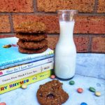 Secret Lentil Cookies on a concrete and brick background. There are some candies scattered around and a glass jug of milk. Some of the cookies are on top of books with the titles on their spines displayed.