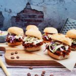 BBQ Coffee Pulled Jackfruit Sliders on parchment paper on a wooden cutting board with coffee beans, a wooden spoon with coffee beans in it, a bowl of coleslaw and a cheese grater on it's side. They are situated on a wooden table surface and have an exposed brick and concrete design on the background wall.