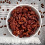 A top-down shot of Cinna Cocoa Coffee-Roasted Nuts in a white bowl on a fancy cloth with whole roasted coffee beans on it situated on a cracked-concrete style countertop.