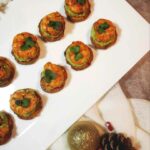 A top-down picture of the Avocado Shrimp Sweet Potato Bites on a white plate. It is on a countertop of white cloth napkins, gold Christmas ornaments plastic cranberries and acorns as a decoration.