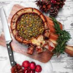 A top-down picture of Baked Cranberry Brie Sourdough Bread Bowl with some bread broken off and cheese oozing out. It is on a wooden paddle board with a bread knife and a sprig of fresh rosemary as a garnish. It is on a white worn wooden background with fake snow, Christmas ornaments and pine cones as decorations.