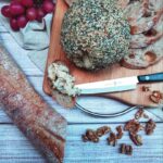 A top-down shot of Everything Bagel Cheeze Ball on a wooden cutting board with a metal handle with slices of baguette and a small serrated knife. It's on a plank wood-style counter top with the rest of the baguette, walnuts, grapes and a beige cloth as decorations surrounding the cutting board.