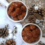 A top-down image of two servings of Honey Garlic Turkey Meatballs in soup bowls on a brown Christmas tablecloth with white snowflakes. Around it are silver and white ornaments and pine cones with fake cotton snow.