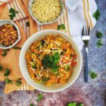 A top-down image of our Protein-Packed Pasta Primavera topped shredded parmesan cheese and basil microgreens on a worn metal-style table. In separate bowls, there is more parmesan cheese and chili flakes. Also on the table is a fork, an orange and white cloth and more microgreens.