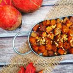 A top-down image of roasted root vegetables in a metal dish with a loop handle. The dish is on a wood table with a wicker cloth, with red autumn leaves and root vegetables as decorations.