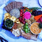 A top-down picture of the Vegan Charcuterie Board. It's on top of a white cloth with a grey-blue pattern on it. It is on a cracked concrete-style counter top. In the top-eight corner, there is a white plate half showing. On the bottom-left corner is another white plate with two small serrated knifes placed on top of each other on it.