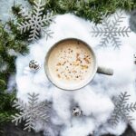 An overhead shot of a latte drink with fake snow, ornaments and evergreen branches as a decoration on a grey concrete-style countertop.