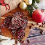 A top-down picture of Chocolate Pomegranate Bark on a cutting board with a whole pomegranate, Christmas ornaments and decorations around it on a dark brown plank wood table.