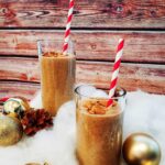 Two smoothie drinks with red and white striped paper straws on a brown wood panel backround. Around it (as decorations) are fake snow, various golden ornaments and decorative pinecones.