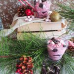 An image of the completed Kids' Candy Cane Trifle recipe in a glass around pink and white mini marshmallows, crushed cookies and Christmas decorations.