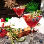 Two red mocktail drinks in cocktail glasses with blueberries and sprigs of rosemary as a garnish. One drink is on a cracked-concrete style countertop and the other is elevated on a wooden cutting board. Scattered around (as decorations) are red, green and gold Christmas ornaments, blueberries, fake snow and a large sprig of rosemary.