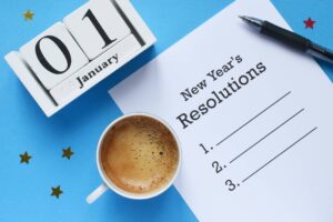 A top-down image of a paper list titled "New Year's Resolutions" with a list of three lines to write your resolutions into. It is surrounded by a pen, a cup of black coffee, gold/red stars, and a white wooden date block set for January 1st. Everything is on a blue background.