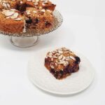 A blueberry square dessert on a white palate with the serving plate that has the rest of the dessert on it on a white background.