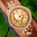 Oatmeal with bananas in it on a wooden cutting board with oats on it as aa decoration. It is in front of a banana leaf background.