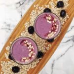 A pink smoothie with outs in it shown from a top-down view. The glasses are on a wooden cutting board with oats and cherries as decorations. The cutting board is on a marble background.