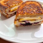 A grilled cheese sandwich on a white plate that's placed on a wooden cutting board with blueberries and mascarpone cheese in tiny bowls as a decoration..