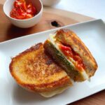 A grilled cheese sandwich on a plate and wooden cutting board with decorative bowls of cherry tomatoes and pesto.