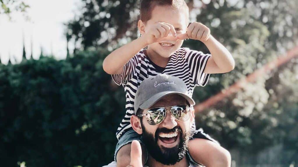 A father with a beard, sunglasses and a hat with his son on his shoulders. They both look happy with joyful expressions.