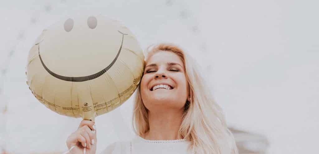 A woman in white smiling with her eyes closed while holding a yellow smiley face balloon beside her head.